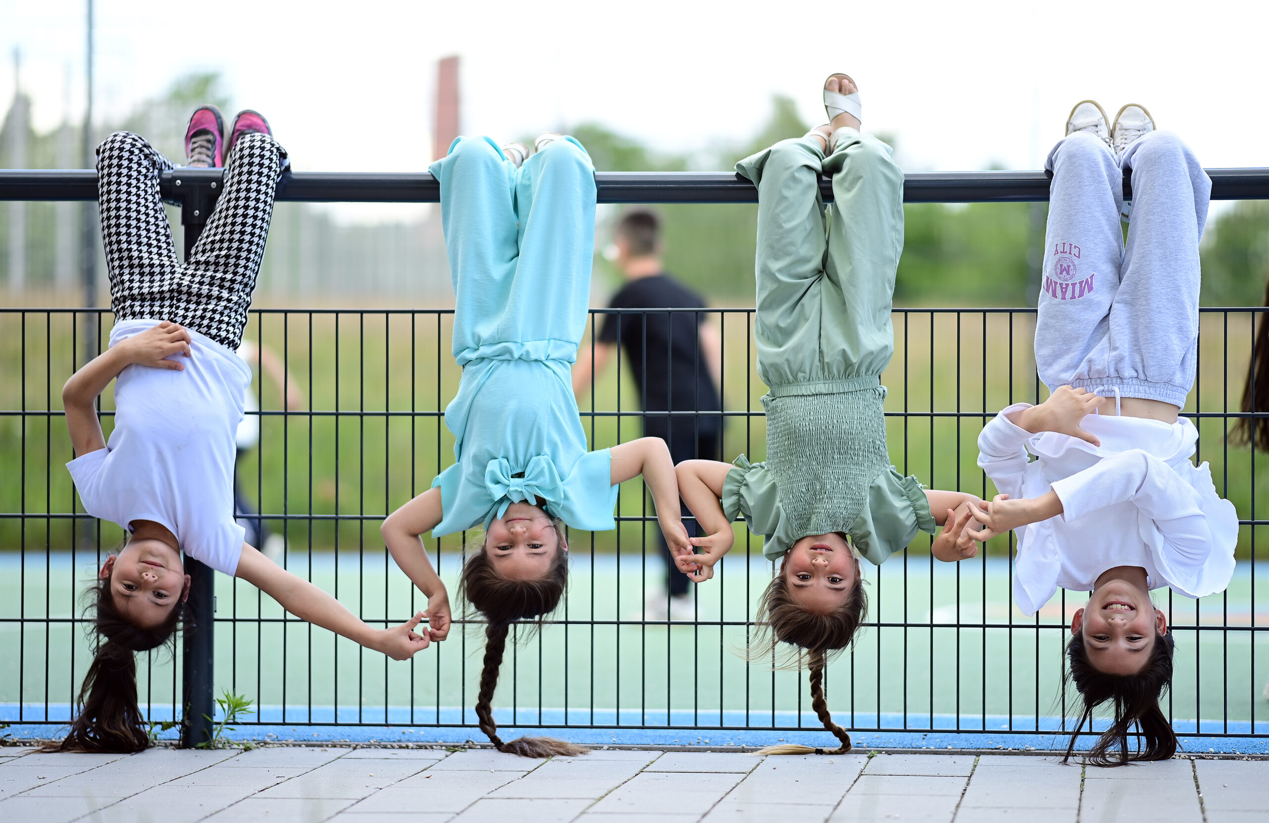 Vier Mädchen hängen an einer Stange und machen den Handstand, im Hintergrund eine Sportfläche und verschwommene Personen.