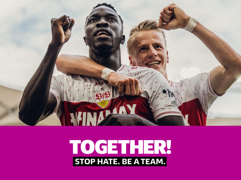 Zwei Fußballspieler, einer mit dunkler Hautfarbe und einer mit heller Hautfarbe, jubeln gemeinsam mit hoch erhobenen Armen. Sie tragen rote Trikots mit dem Schriftzug "SVS". Darunter steht in großen Buchstaben "TOGETHER!" und darunter kleiner "STOP HATE. BE A TEAM.".