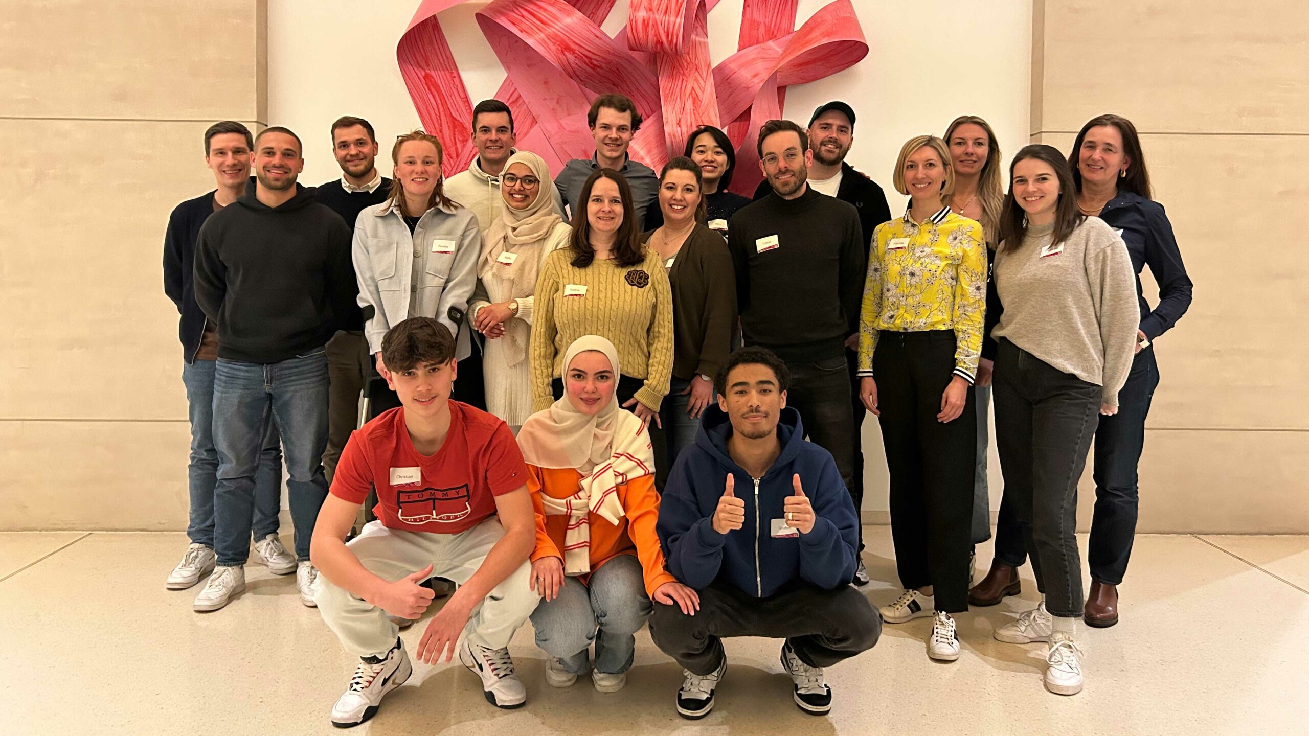 Eine Gruppe von jungen Erwachsenen posiert für ein Gruppenfoto in einem hellen Raum. Sie lächeln in die Kamera und einige zeigen den Daumen hoch. Die Personen tragen vielfältige, legere Kleidung. Im Hintergrund ist eine abstrakte, rosa Skulptur und eine helle Wand zu sehen.