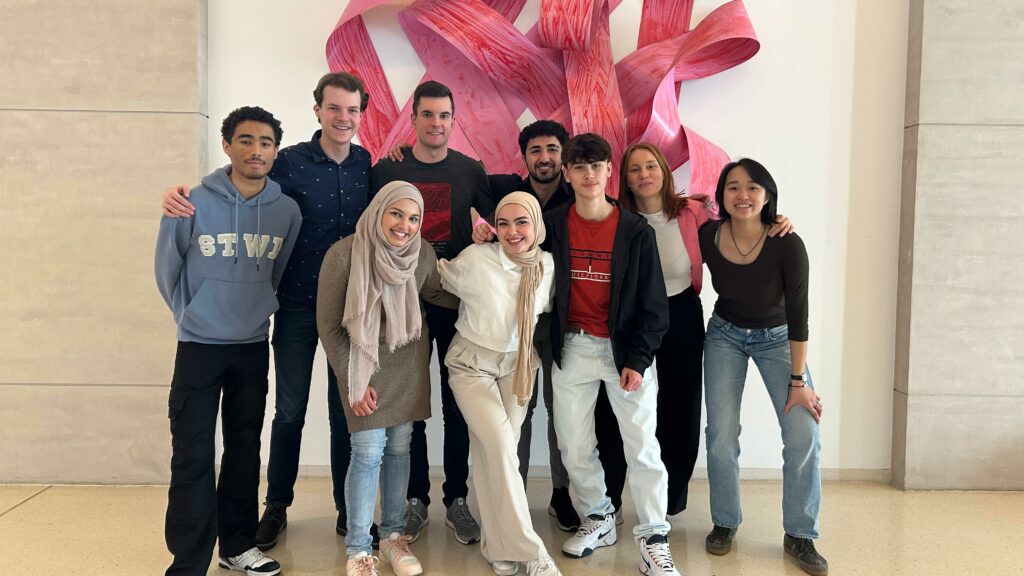 Eine Gruppe von acht jungen Menschen steht lächelnd vor einer abstrakten, roten Wandinstallation. Sie sind vielfältig gekleidet und wirken wie ein Team.