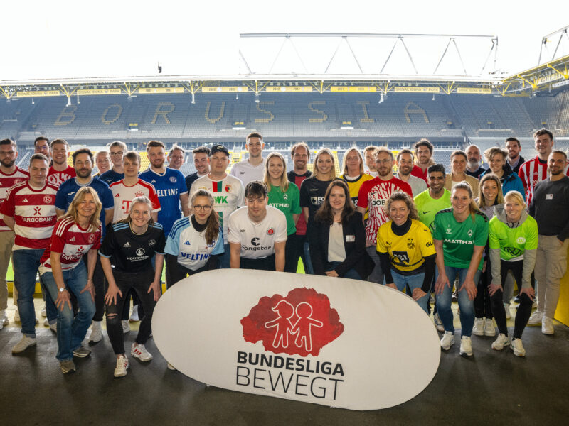 Eine große Gruppe von Menschen steht im Signal Iduna Park in Dortmund vor der Logo-Wand "Bundesliga bewegt" der DFL Stiftung. Viele tragen Trainingsanzüge oder Trikots verschiedener Bundesligavereine. Es handelt sich um ein Netzwerktreffen der DFL Stiftung.