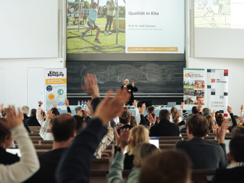Ein Mann steht auf einer Bühne vor einer großen Gruppe von Menschen, die alle die Hände gehoben haben. Im Hintergrund ist eine Präsentation mit dem Titel "Qualität in Kitas" und dem Namen "Prof. Dr. Rolf Schwarz" zu sehen. Das Bild zeigt eine lebhafte Interaktion und möglicherweise eine Veranstaltung oder einen Workshop der DFL Stiftung.