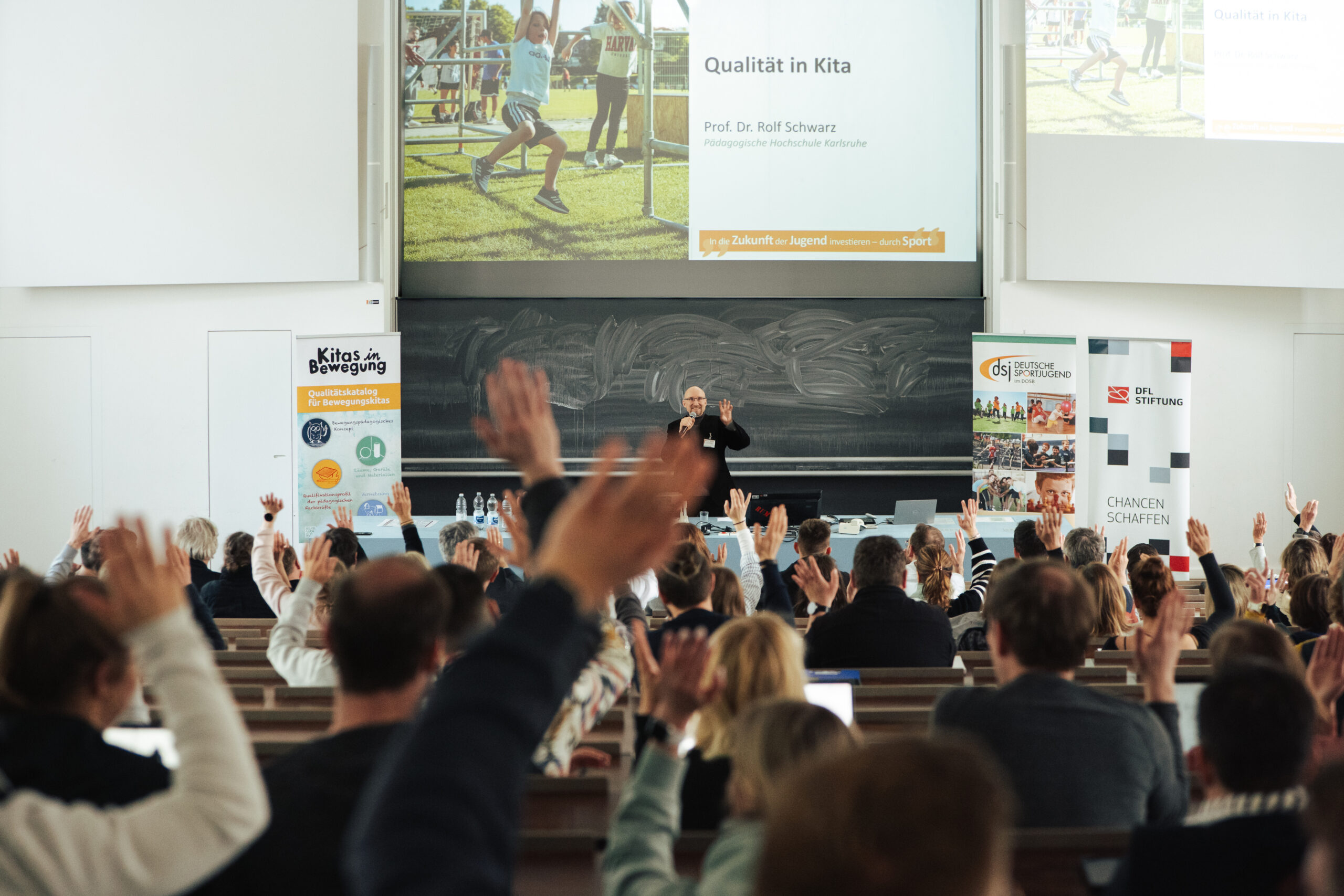 Ein Mann steht auf einer Bühne vor einer großen Gruppe von Menschen, die alle die Hände gehoben haben. Im Hintergrund ist eine Präsentation mit dem Titel "Qualität in Kitas" und dem Namen "Prof. Dr. Rolf Schwarz" zu sehen. Das Bild zeigt eine lebhafte Interaktion und möglicherweise eine Veranstaltung oder einen Workshop der DFL Stiftung.