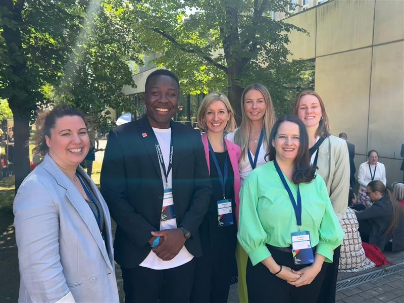 Eine Gruppe von lächelnden Personen steht für ein gemeinsames Foto zusammen. Die Gruppe besteht aus verschiedenen Personen, darunter Frauen und Männer, die professionell gekleidet sind und Lanyards mit Ausweisen tragen. Im Hintergrund sind Bäume und ein Gebäude zu sehen.