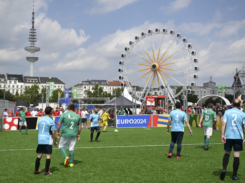 Fußballspiel in der Fan-Zone bei der UEFA EURO 2024 mit dem Fernsehturm im Hintergrund. Mehrere Spieler in hellblauen und grünen Trikots sind auf dem Rasen zu sehen, während im Hintergrund ein Riesenrad und Zelte stehen.