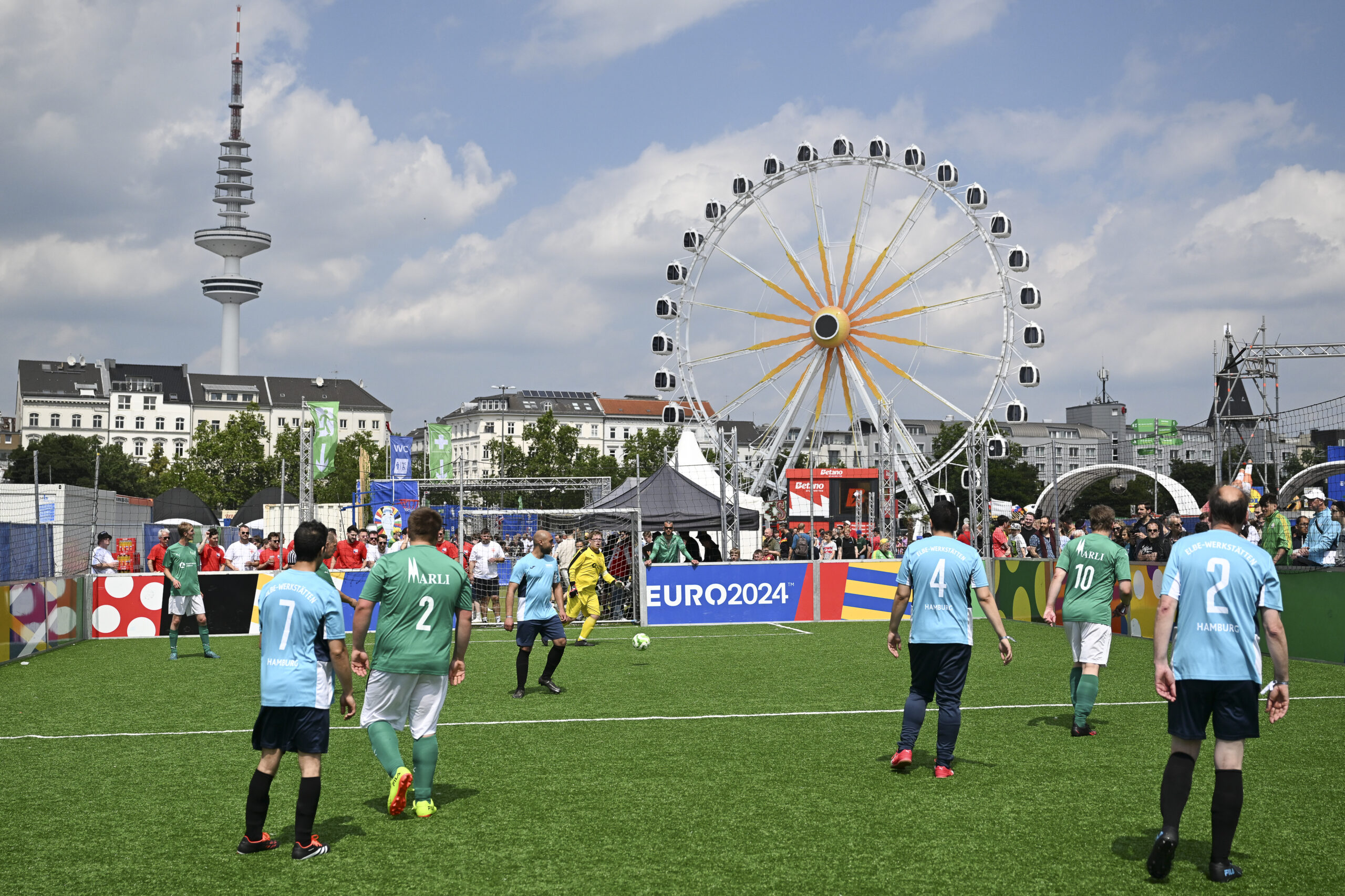 Fußballspiel in der Fan-Zone bei der UEFA EURO 2024 mit dem Fernsehturm im Hintergrund. Mehrere Spieler in hellblauen und grünen Trikots sind auf dem Rasen zu sehen, während im Hintergrund ein Riesenrad und Zelte stehen.