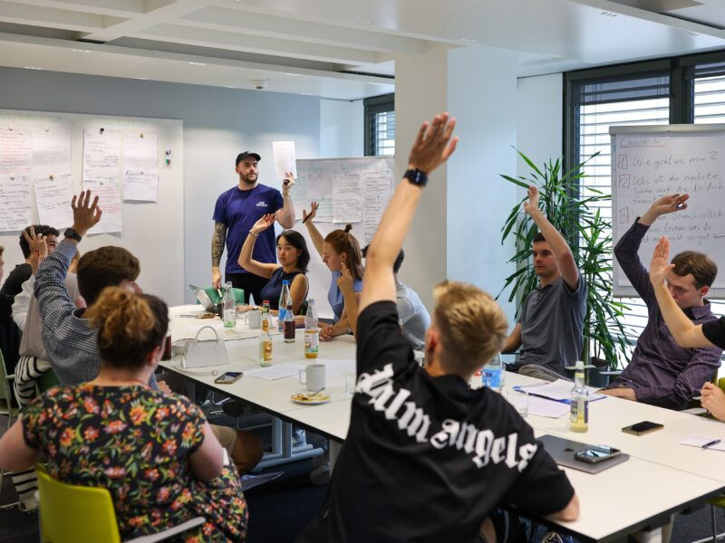 Jugendliche sitzen an einem Konferenztisch in einem hellen Raum und heben die Hände, während eine Vortragende im Hintergrund steht. Im Fokus steht das Treffen des Jugendgremiums der DFL Stiftung in Frankfurt.