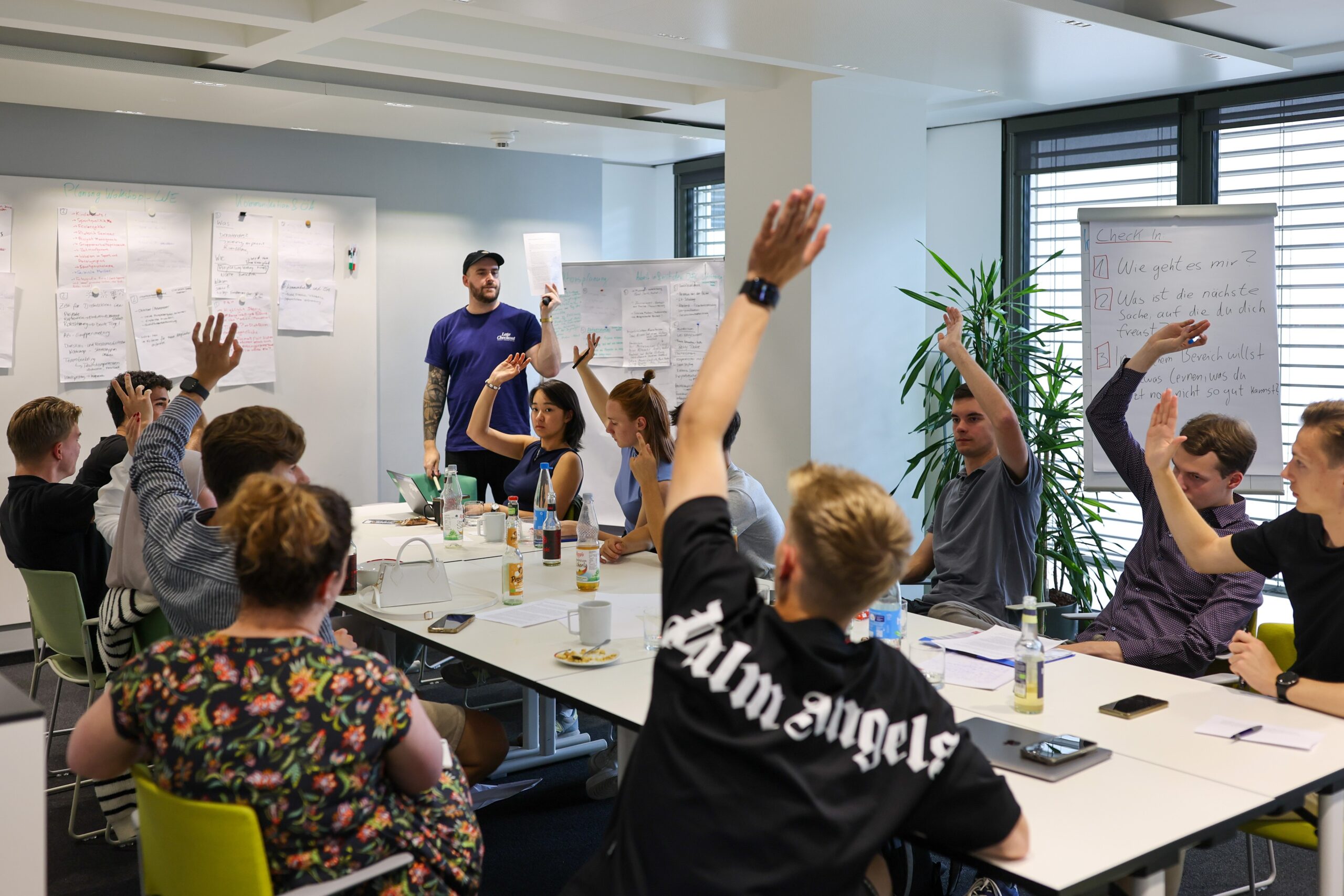 Jugendliche sitzen an einem Konferenztisch in einem hellen Raum und heben die Hände, während eine Vortragende im Hintergrund steht. Im Fokus steht das Treffen des Jugendgremiums der DFL Stiftung in Frankfurt.