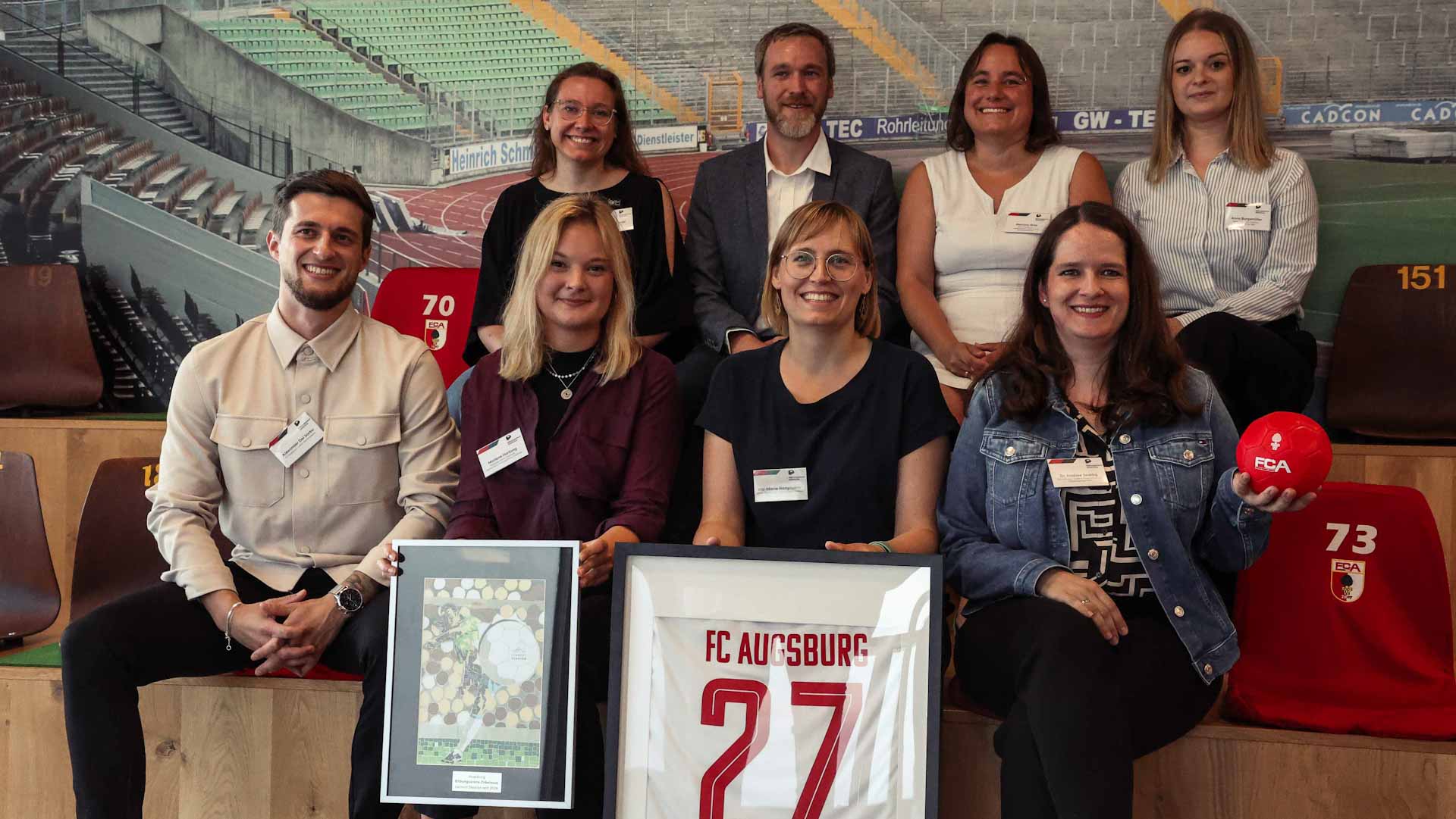 Eine Gruppe von etwa zehn Personen sitzt oder steht lächelnd vor einer Wand mit Stadionmotiven. Im Mittelpunkt hält eine Frau ein gerahmtes Foto, auf dem vermutlich eine Sportanlage zu sehen ist. Alle tragen Kleidung mit dem Logo des FC Augsburg oder Namensschilder, was auf ein gemeinsames Projekt oder eine Veranstaltung im Rahmen der DFL Stiftung hindeutet.