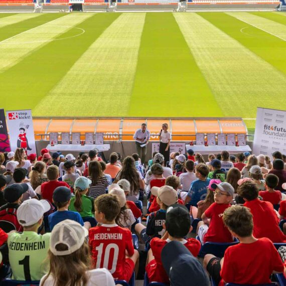 Siegerehrung des "step kickt!"-Projekts der DFL Stiftung im Fußballstadion mit vielen Kindern und Jugendlichen in roten Trikots, die auf die Bühne und einen Redner blicken. Im Hintergrund Banner mit dem Logo "fit4future foundation".
