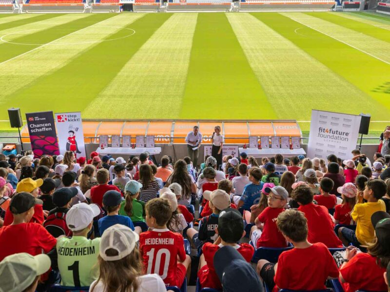 Siegerehrung des "step kickt!"-Projekts der DFL Stiftung im Fußballstadion mit vielen Kindern und Jugendlichen in roten Trikots, die auf die Bühne und einen Redner blicken. Im Hintergrund Banner mit dem Logo "fit4future foundation".