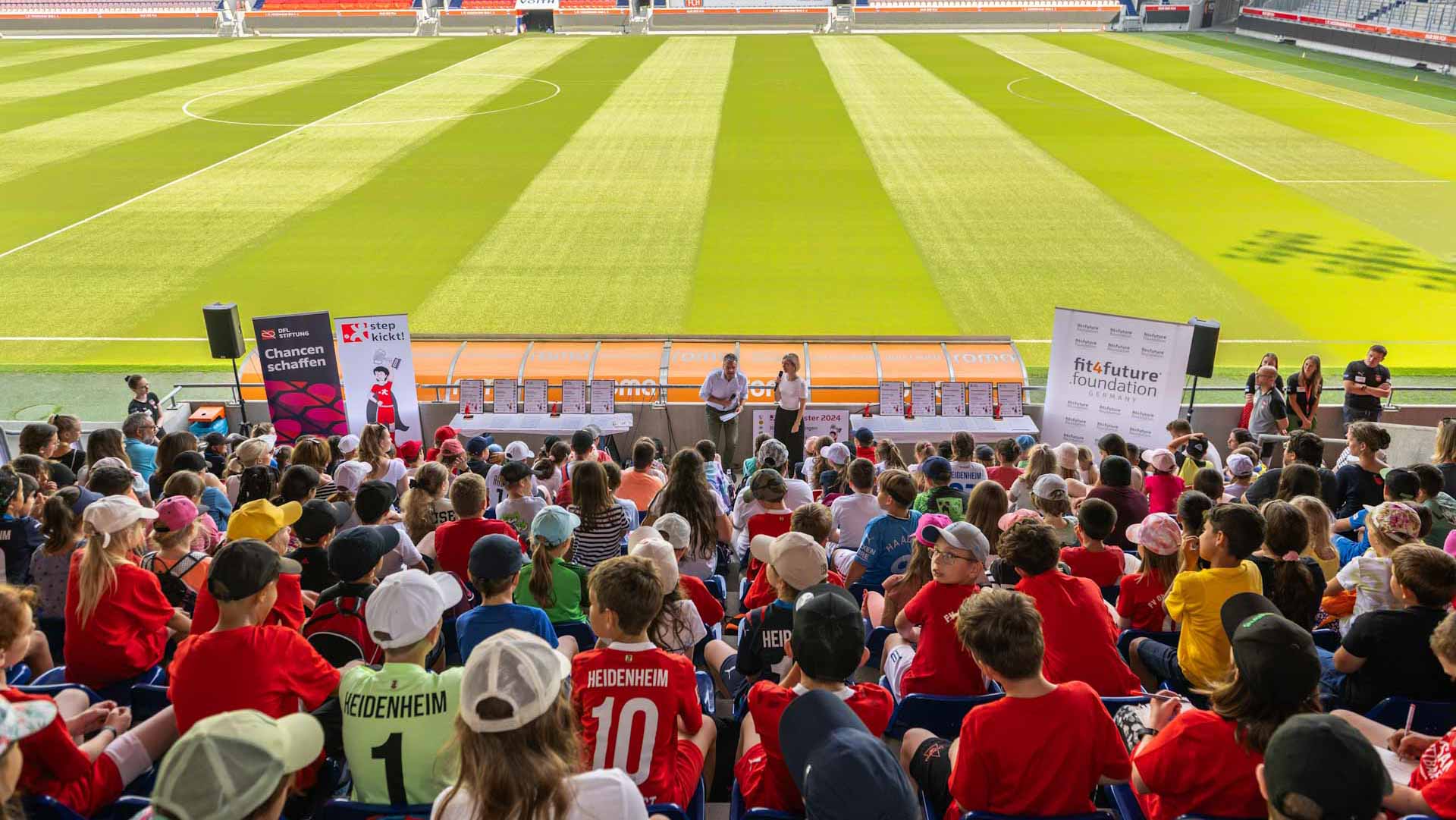 Siegerehrung des "step kickt!"-Projekts der DFL Stiftung im Fußballstadion mit vielen Kindern und Jugendlichen in roten Trikots, die auf die Bühne und einen Redner blicken. Im Hintergrund Banner mit dem Logo "fit4future foundation".