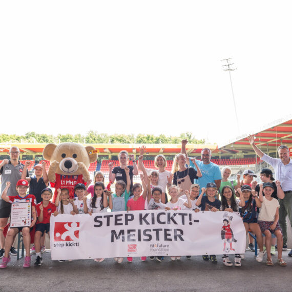 Eine Gruppe von Kindern und Jugendlichen steht vor einer Fußballarena und hält ein Banner mit der Aufschrift "STEP KICKT! MEISTER". Einige halten Pokale hoch und lächeln in die Kamera. Im Hintergrund ist das Stadion zu sehen und Banner der DFL Stiftung und des Projekts "step kickt!".
