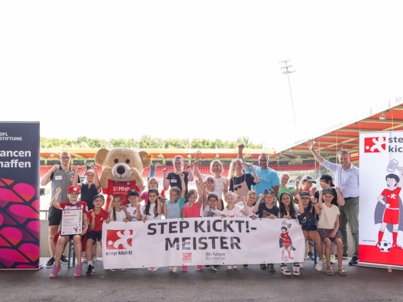 Eine Gruppe von Kindern und Jugendlichen steht vor einer Fußballarena und hält ein Banner mit der Aufschrift "STEP KICKT! MEISTER". Einige halten Pokale hoch und lächeln in die Kamera. Im Hintergrund ist das Stadion zu sehen und Banner der DFL Stiftung und des Projekts "step kickt!".