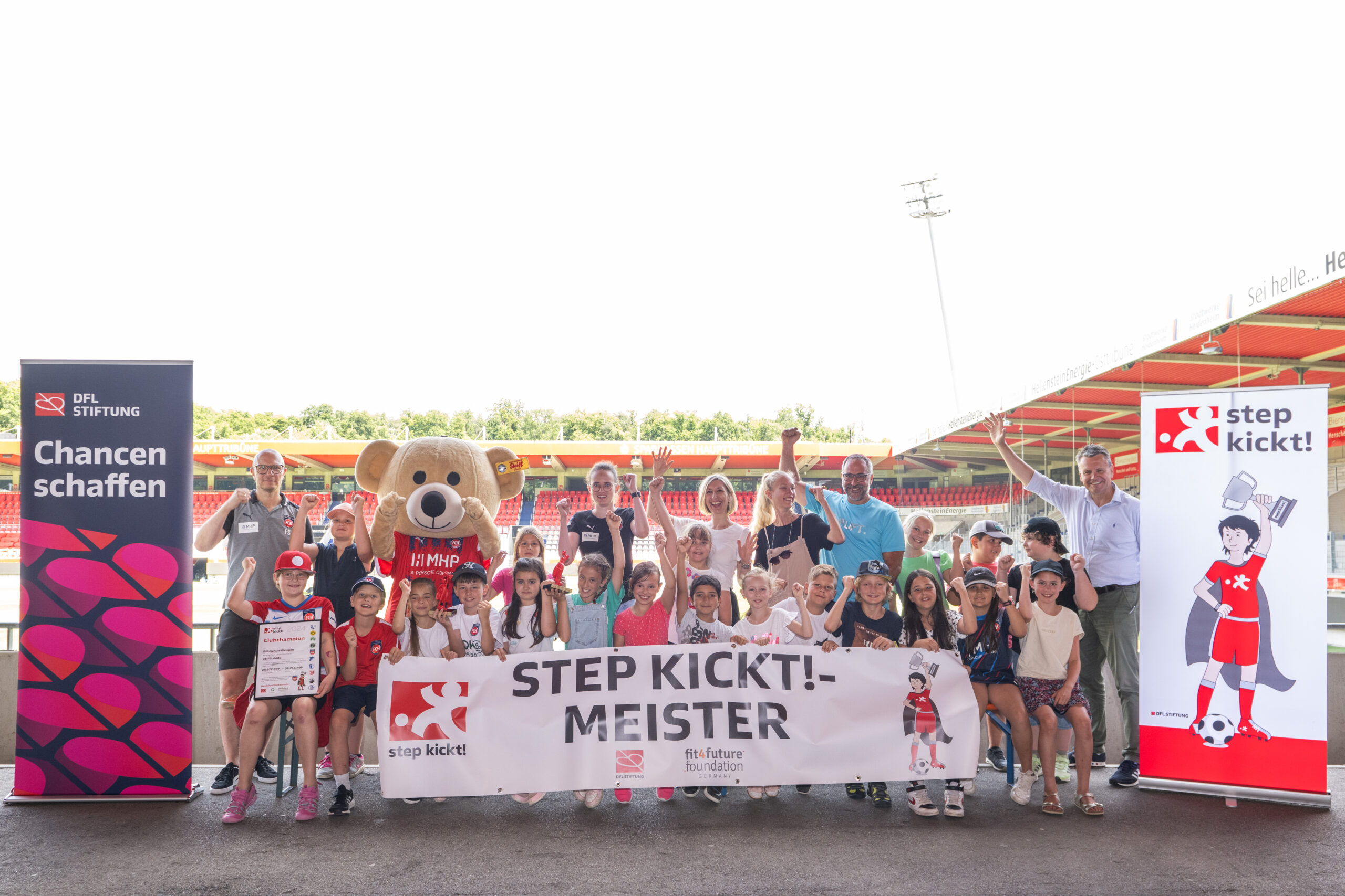 Eine Gruppe von Kindern und Jugendlichen steht vor einer Fußballarena und hält ein Banner mit der Aufschrift "STEP KICKT! MEISTER". Einige halten Pokale hoch und lächeln in die Kamera. Im Hintergrund ist das Stadion zu sehen und Banner der DFL Stiftung und des Projekts "step kickt!".