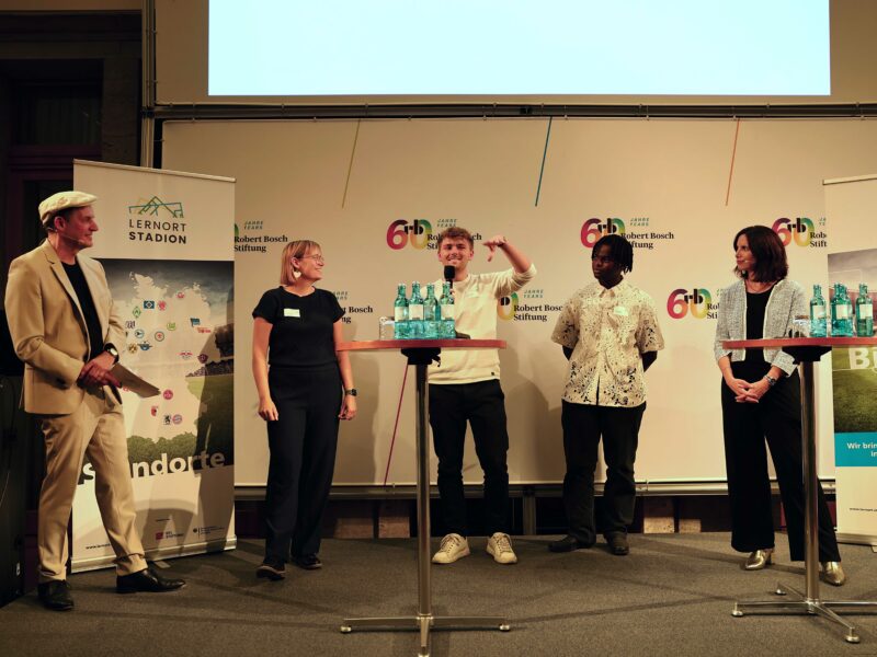 Eine Gruppe von sechs Personen steht auf einer Bühne vor einer Wand mit Logos und Bannern zur "DFL Stiftung" und "15 Jahre Lernort Stadion". Die Personen halten jeweils ein großes, gerahmtes Bild vor sich. Im Hintergrund ist ein rotes Banner mit der Aufschrift "Bildung verändert Zukunft" zu sehen.