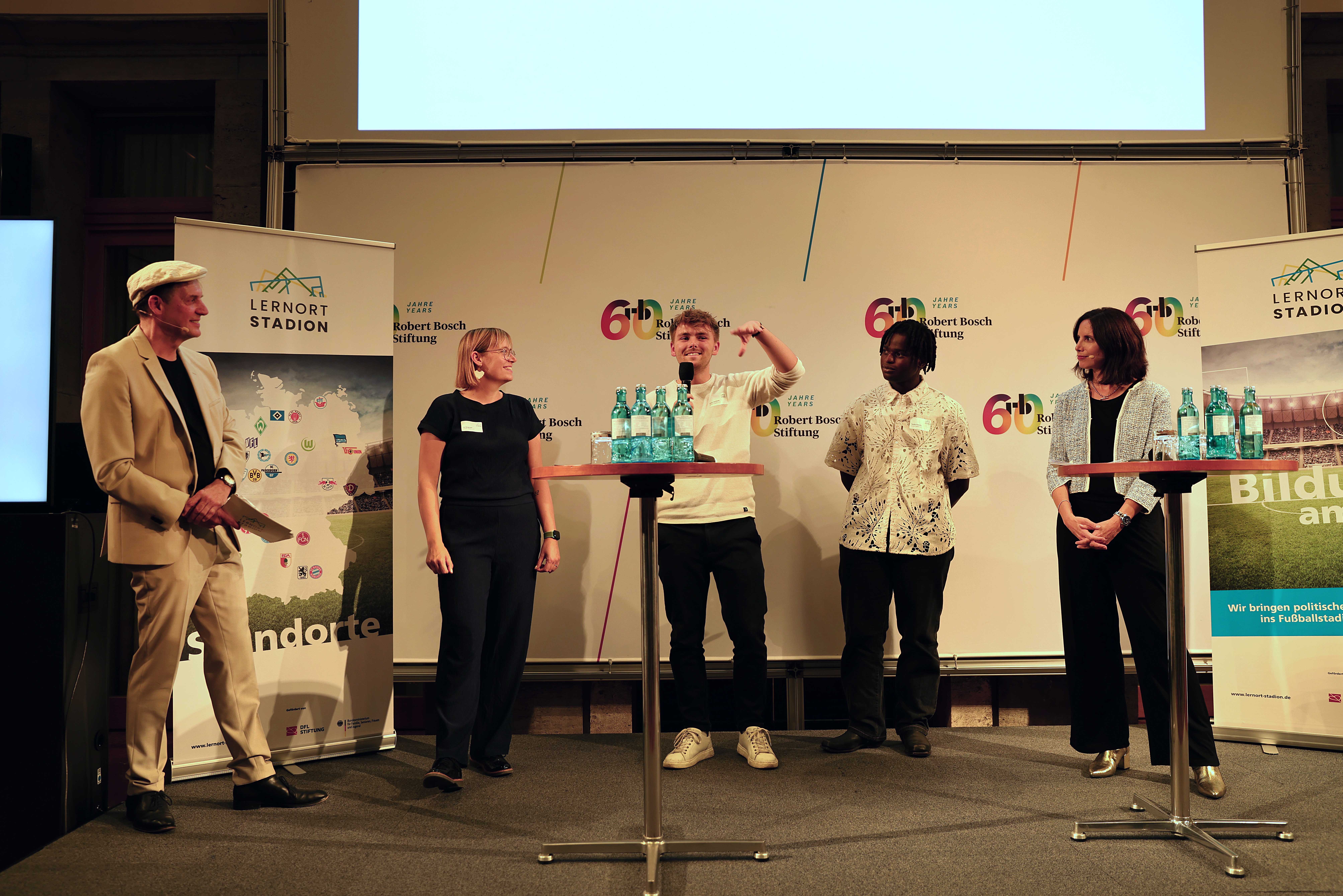 Eine Gruppe von sechs Personen steht auf einer Bühne vor einer Wand mit Logos und Bannern zur "DFL Stiftung" und "15 Jahre Lernort Stadion". Die Personen halten jeweils ein großes, gerahmtes Bild vor sich. Im Hintergrund ist ein rotes Banner mit der Aufschrift "Bildung verändert Zukunft" zu sehen.