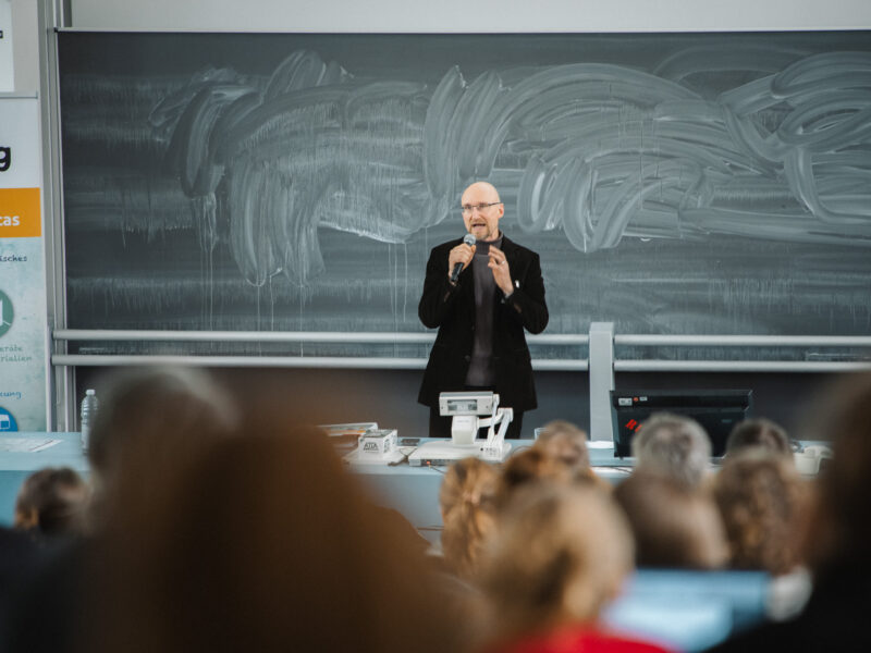 Ein Mann hält einen Vortrag vor einer Gruppe von Teilnehmenden. Im Hintergrund eine grüne Tafel und Plakate mit Schriftzügen.