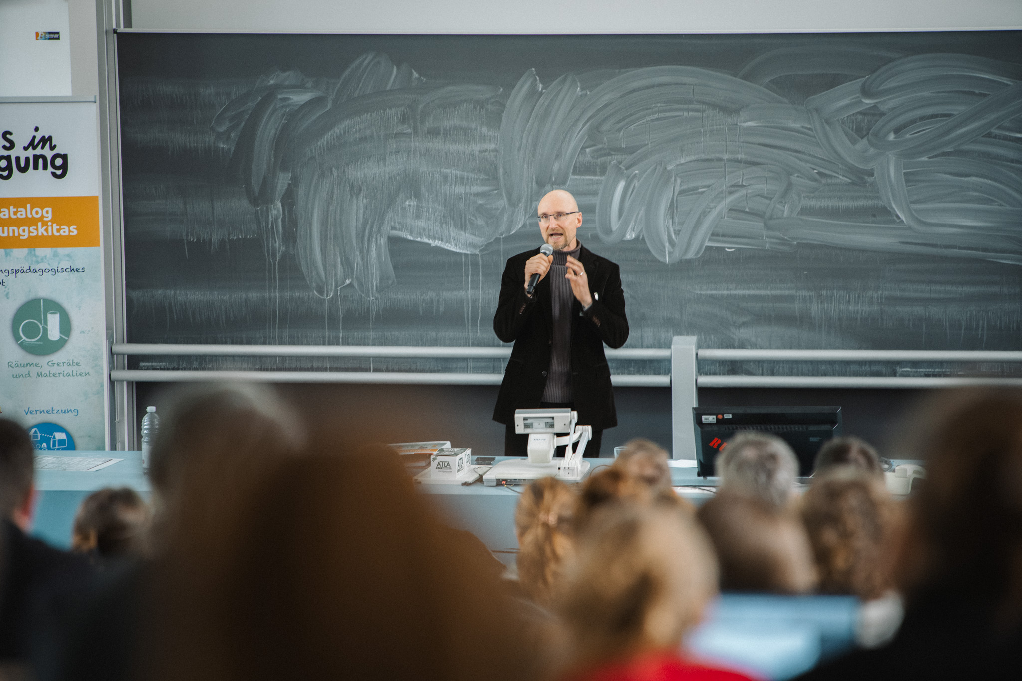 Ein Mann hält einen Vortrag vor einer Gruppe von Teilnehmenden. Im Hintergrund eine grüne Tafel und Plakate mit Schriftzügen.