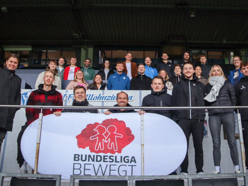 Eine Gruppe von Menschen steht auf einer Tribüne eines Fußballstadions und hält ein Banner mit dem Aufdruck "BUNDESLIGA BEWEGT" und einem roten Symbol, das stilisierte Figuren von Erwachsenen und Kindern darstellt.