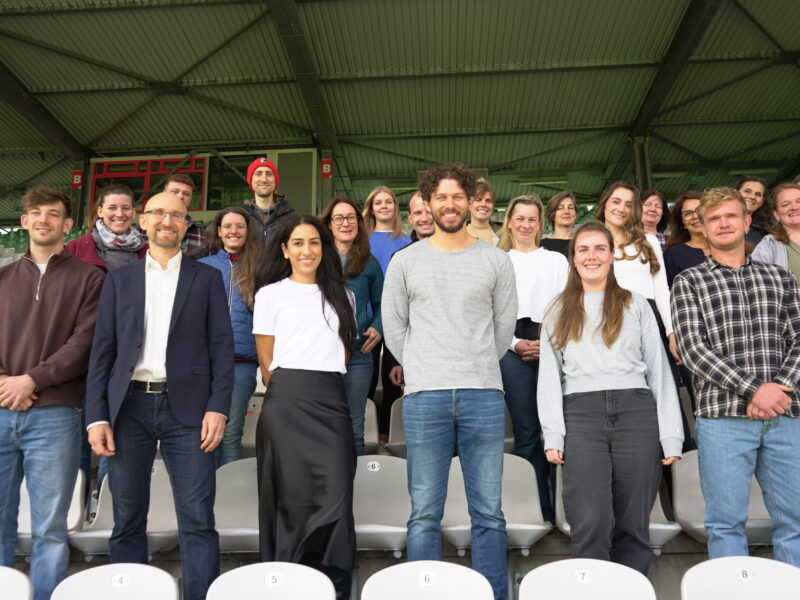 Eine Gruppe von etwa 20 Personen steht auf den Sitzen einer Stadiontribüne und lächelt in die Kamera. Die Personen sind vielfältig in Alter und Geschlecht und tragen verschiedene Kleidungsstile. Im Hintergrund ist ein Teil der Stadionkonstruktion zu sehen.