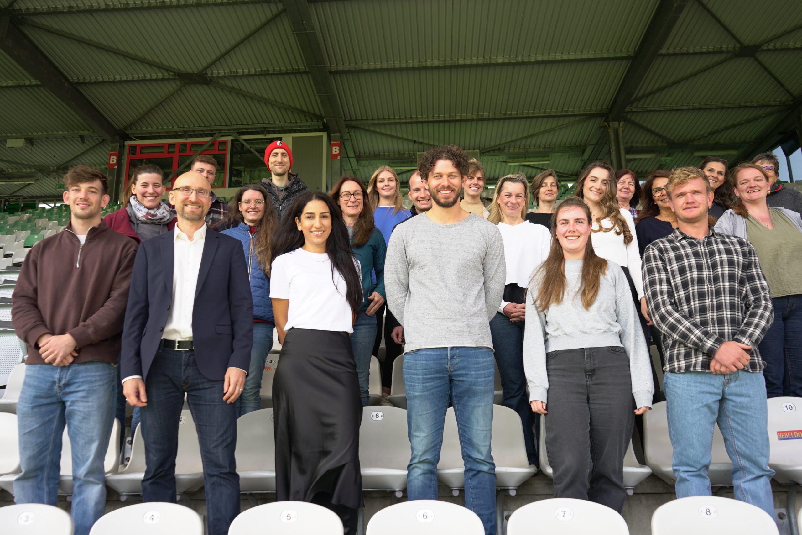 Eine Gruppe von etwa 20 Personen steht auf den Sitzen einer Stadiontribüne und lächelt in die Kamera. Die Personen sind vielfältig in Alter und Geschlecht und tragen verschiedene Kleidungsstile. Im Hintergrund ist ein Teil der Stadionkonstruktion zu sehen.