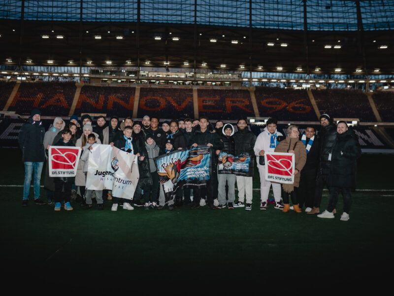 Eine Gruppe von Menschen steht auf dem Rasen eines Fußballstadions. Einige halten Banner mit dem Logo der DFL Stiftung und dem Schriftzug "Jugendzentrum" hoch. Im Hintergrund sind die leeren Zuschauertribünen zu sehen.