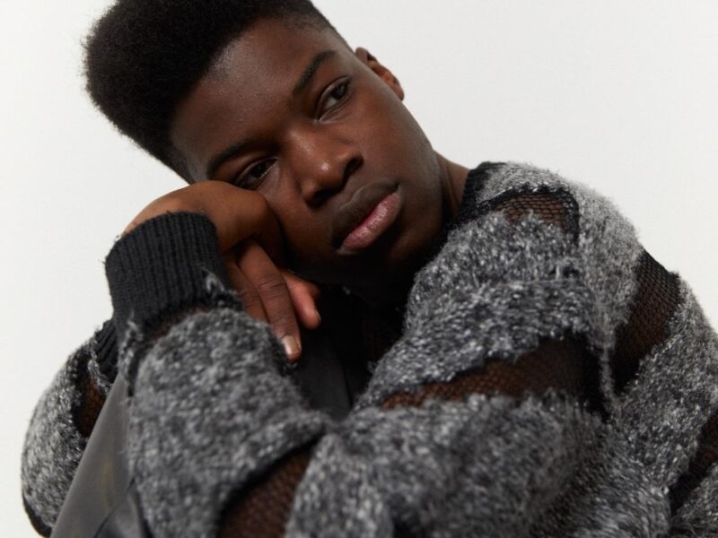 Ein junger Mann mit dunkler Hautfarbe blickt nachdenklich in die Kamera. Er trägt einen grauen Strickpullover und eine schwarze Lederjacke. Das Porträt vermittelt einen Eindruck von Verletzlichkeit und innerer Zerrissenheit.