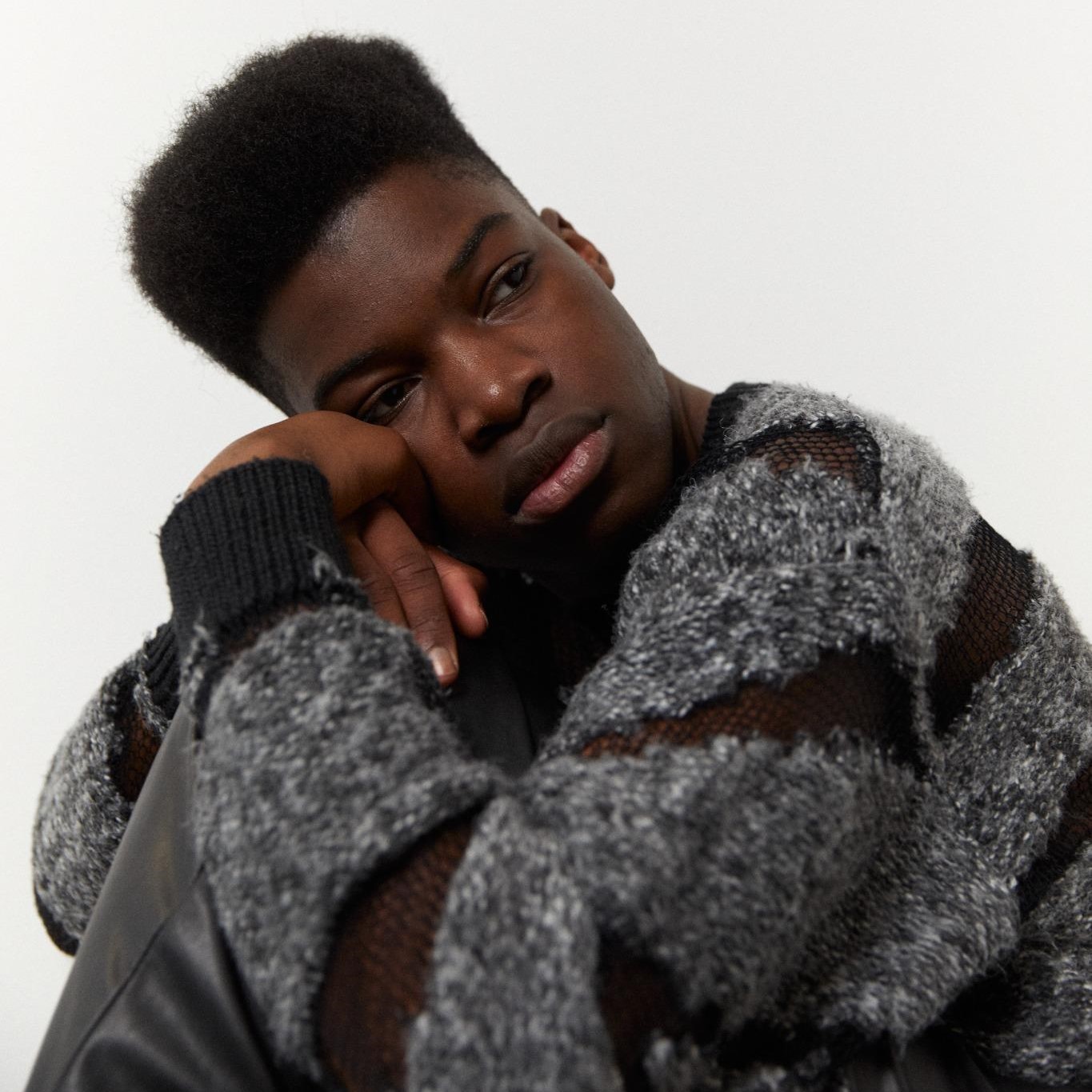 Ein junger Mann mit dunkler Hautfarbe blickt nachdenklich in die Kamera. Er trägt einen grauen Strickpullover und eine schwarze Lederjacke. Das Porträt vermittelt einen Eindruck von Verletzlichkeit und innerer Zerrissenheit.