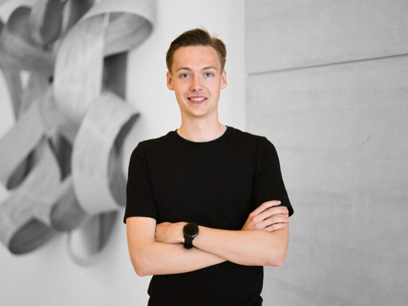 Junge Person mit verschränkten Armen, lächelnd, im Hintergrund eine abstrakte Wandinstallation – Teilnehmer des Treffens des Jugendgremiums der DFL Stiftung.