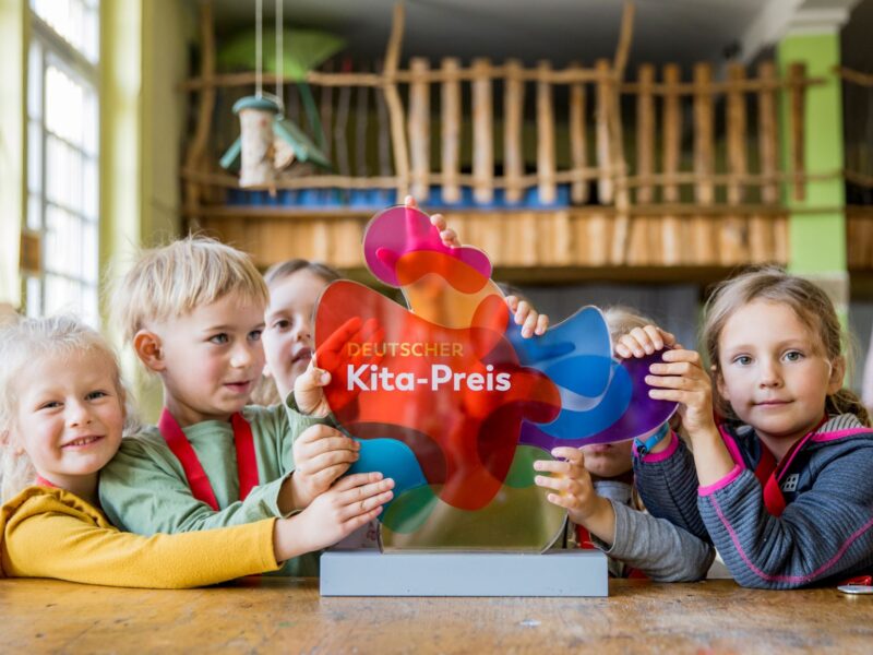 Eine Gruppe von Kindern steht erwartungsvoll vor einer farbenfrohen Trophäe mit der Aufschrift "Deutscher Kita-Preis".