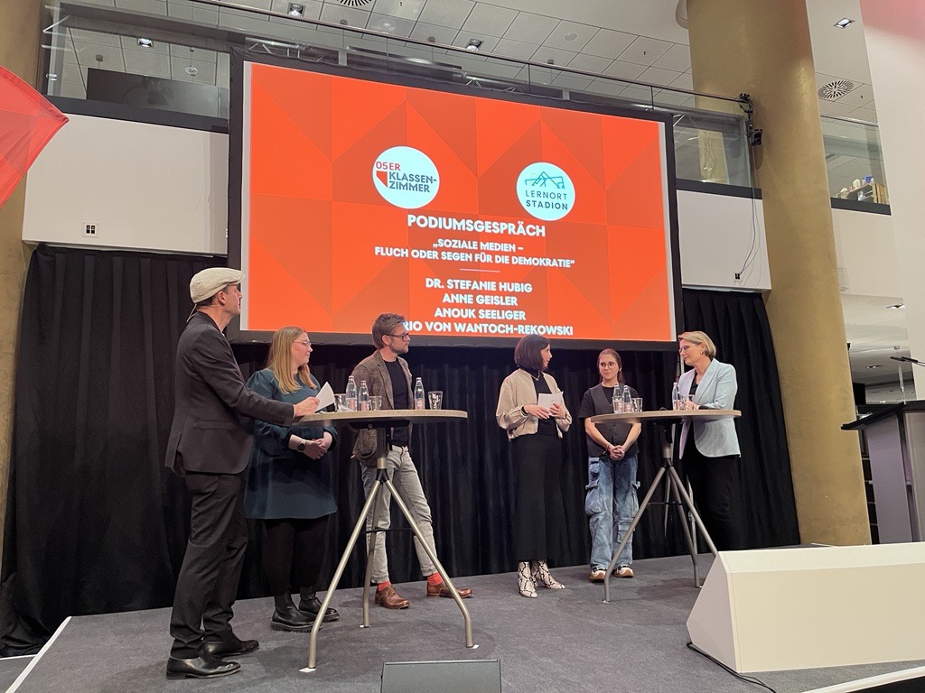 Eine Podiumsdiskussion mit vier Personen auf einer Bühne. Im Hintergrund eine Leinwand mit dem Titel "SOZIALE MEDIEN – FLUCH ODER SEGEN FÜR DIE DEMOKRATIE?". Von links sind ein Mann im Anzug, eine Frau in einem beigen Blazer, eine Frau mit Jeans und ein Mann zu sehen.