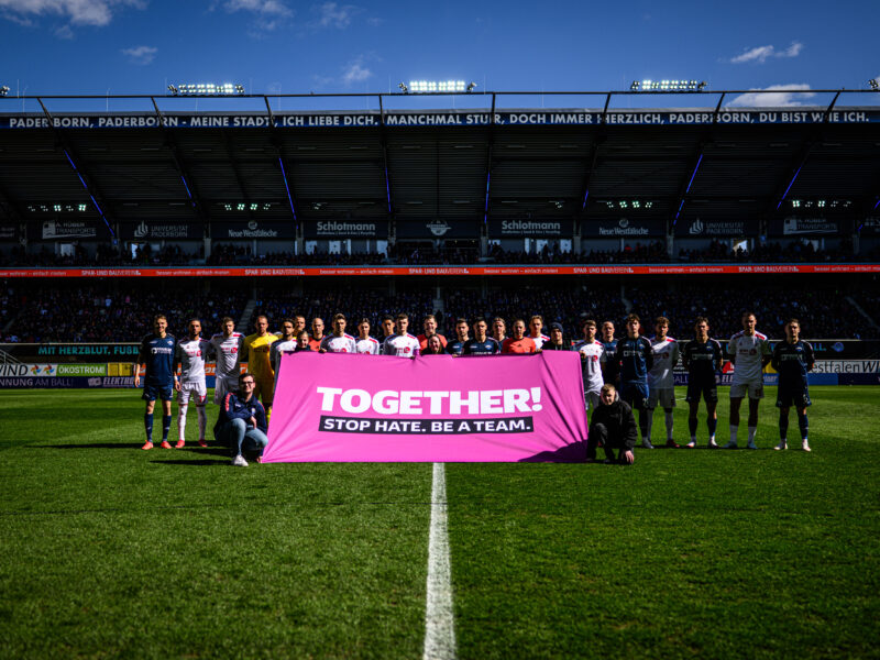 Spieler beider Mannschaften, SC Paderborn 07 und 1. FC Köln, stehen gemeinsam mit einem Banner im Stadion, auf dem "TOGETHER. STOP HATE." steht. Im Hintergrund sind die Zuschauerränge zu sehen.