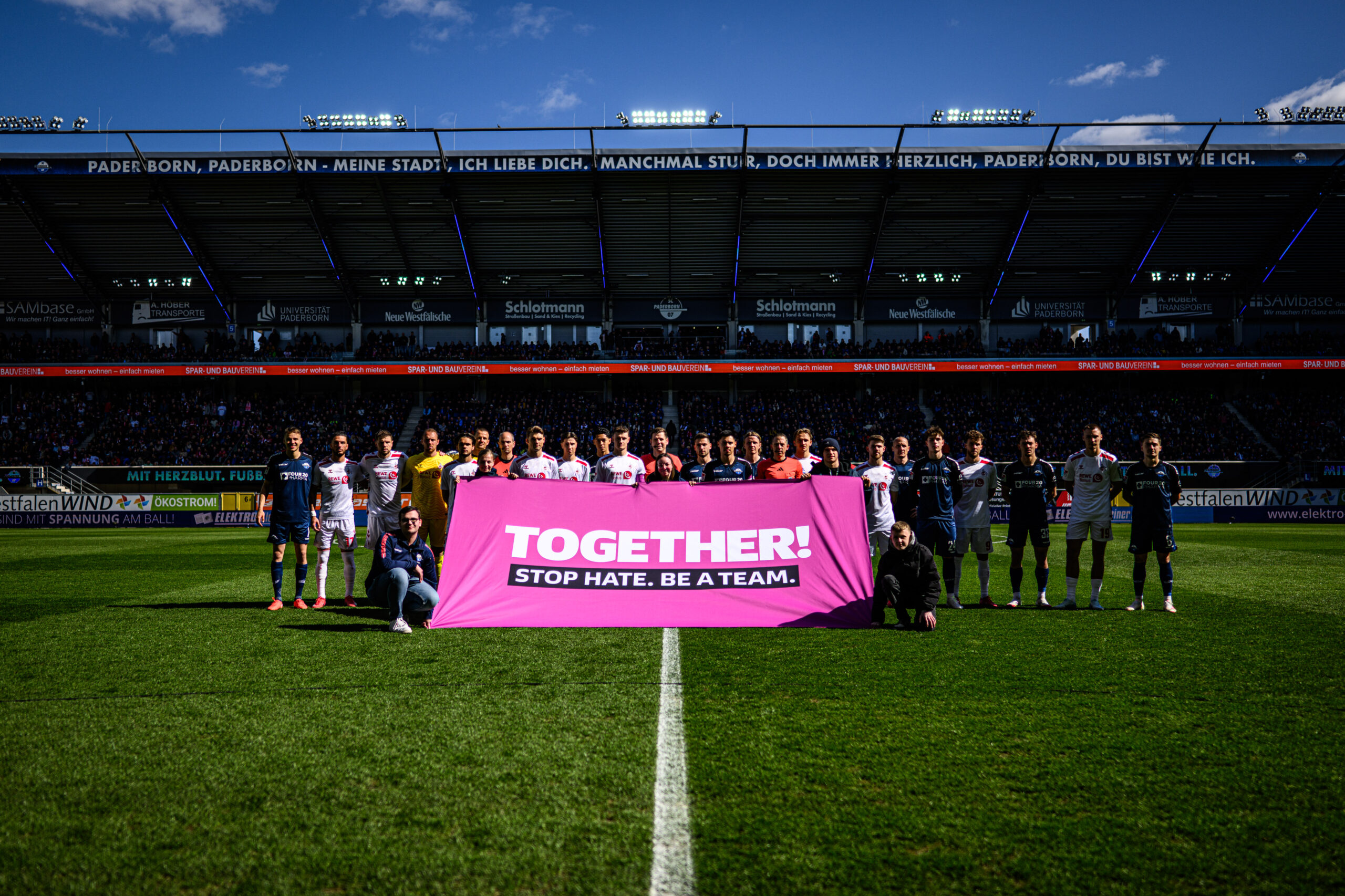 Spieler beider Mannschaften, SC Paderborn 07 und 1. FC Köln, stehen gemeinsam mit einem Banner im Stadion, auf dem "TOGETHER. STOP HATE." steht. Im Hintergrund sind die Zuschauerränge zu sehen.