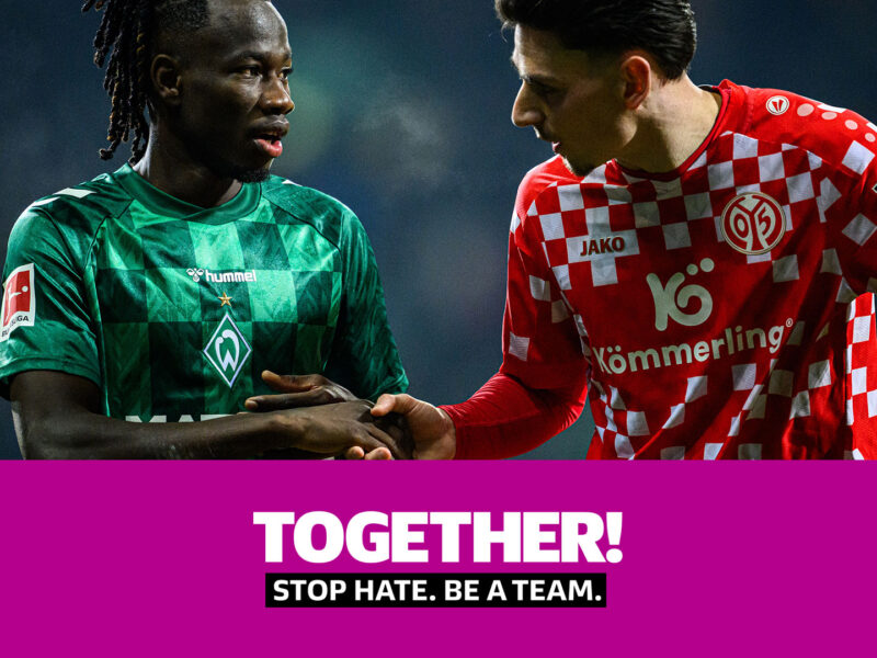 Zwei Fußballspieler, einer mit grünem Trikot des SV Werder Bremen und einer mit rotem Trikot des 1. FSV Mainz 05, reichen sich die Hände. Darunter steht der Slogan "TOGETHER. STOP HATE. BE A TEAM." auf lila Hintergrund.