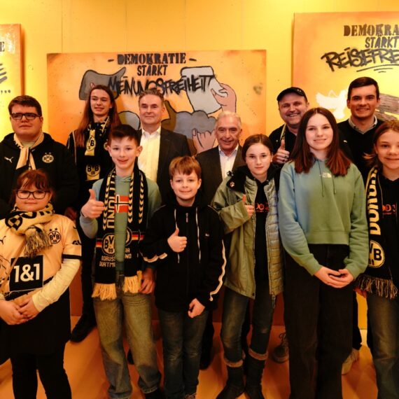 Eine Gruppe von Kindern und Jugendlichen steht vor einer Wand mit Graffiti-Schriftzügen, die "Demokratie stärkt" und "Pressefreiheit" zeigen. Viele der Jugendlichen halten Daumen hoch und tragen Borussia Dortmund Fanartikel.