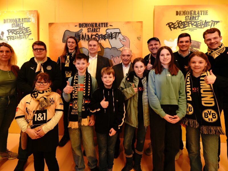Eine Gruppe von Kindern und Jugendlichen steht vor einer Wand mit Graffiti-Schriftzügen, die "Demokratie stärkt" und "Pressefreiheit" zeigen. Viele der Jugendlichen halten Daumen hoch und tragen Borussia Dortmund Fanartikel.