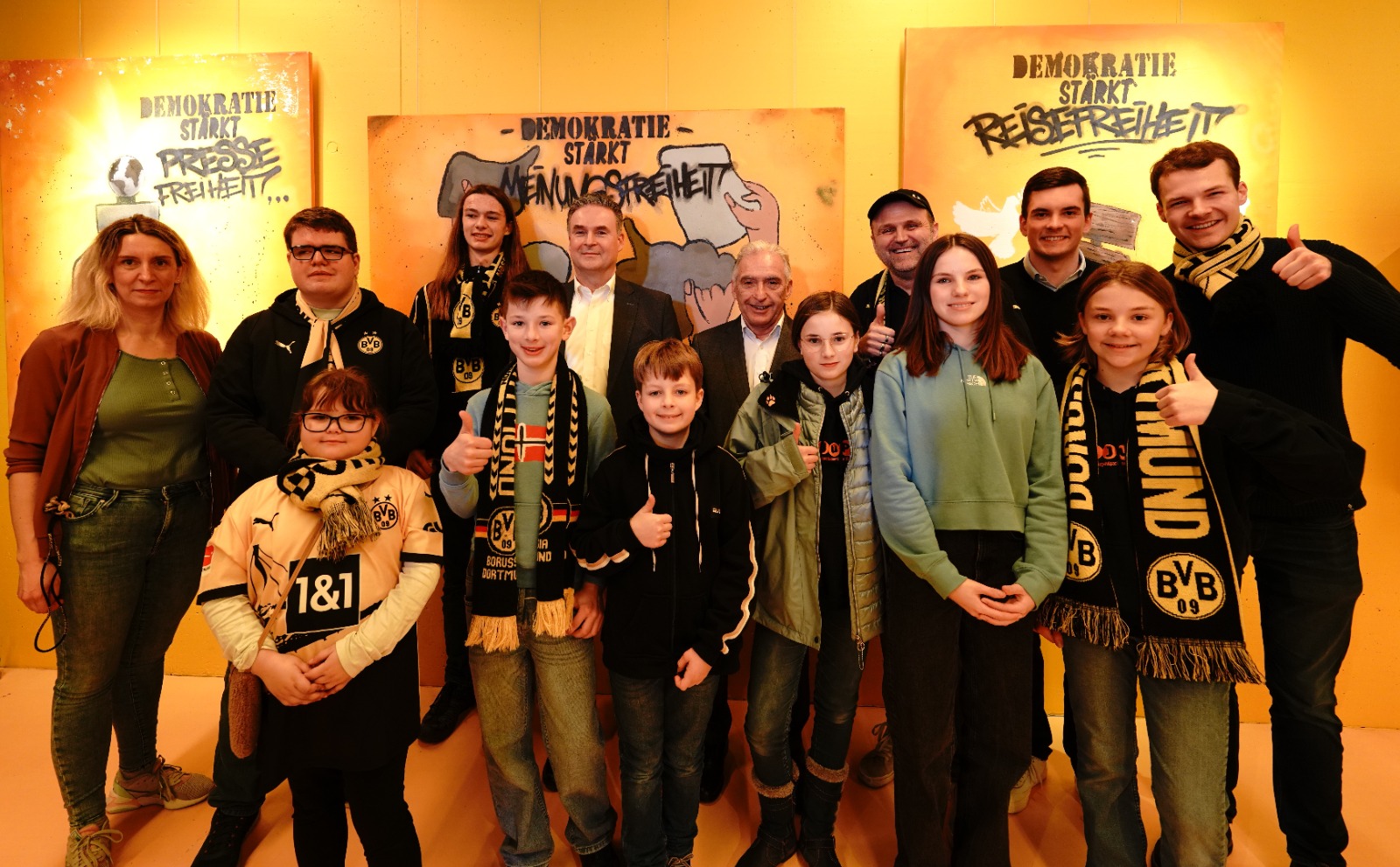 Eine Gruppe von Kindern und Jugendlichen steht vor einer Wand mit Graffiti-Schriftzügen, die "Demokratie stärkt" und "Pressefreiheit" zeigen. Viele der Jugendlichen halten Daumen hoch und tragen Borussia Dortmund Fanartikel.