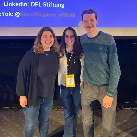 Drei lächelnde Personen stehen vor einer blauen Wand mit den Logos von LinkedIn und TikTok. Die Person links trägt ein dunkelblaues Hemd, die mittlere Person ein rotes Hemd und eine Lanyard mit einer Namenskarte, und die Person rechts ein blaues Hemd mit einem grünen Pullover darüber.