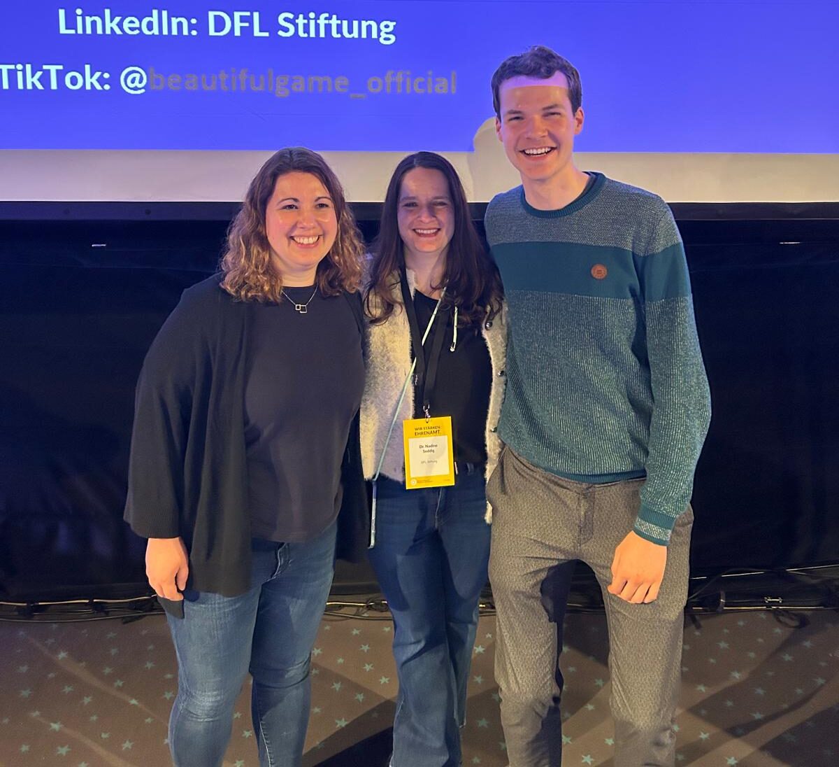 Drei lächelnde Personen stehen vor einer blauen Wand mit den Logos von LinkedIn und TikTok. Die Person links trägt ein dunkelblaues Hemd, die mittlere Person ein rotes Hemd und eine Lanyard mit einer Namenskarte, und die Person rechts ein blaues Hemd mit einem grünen Pullover darüber.