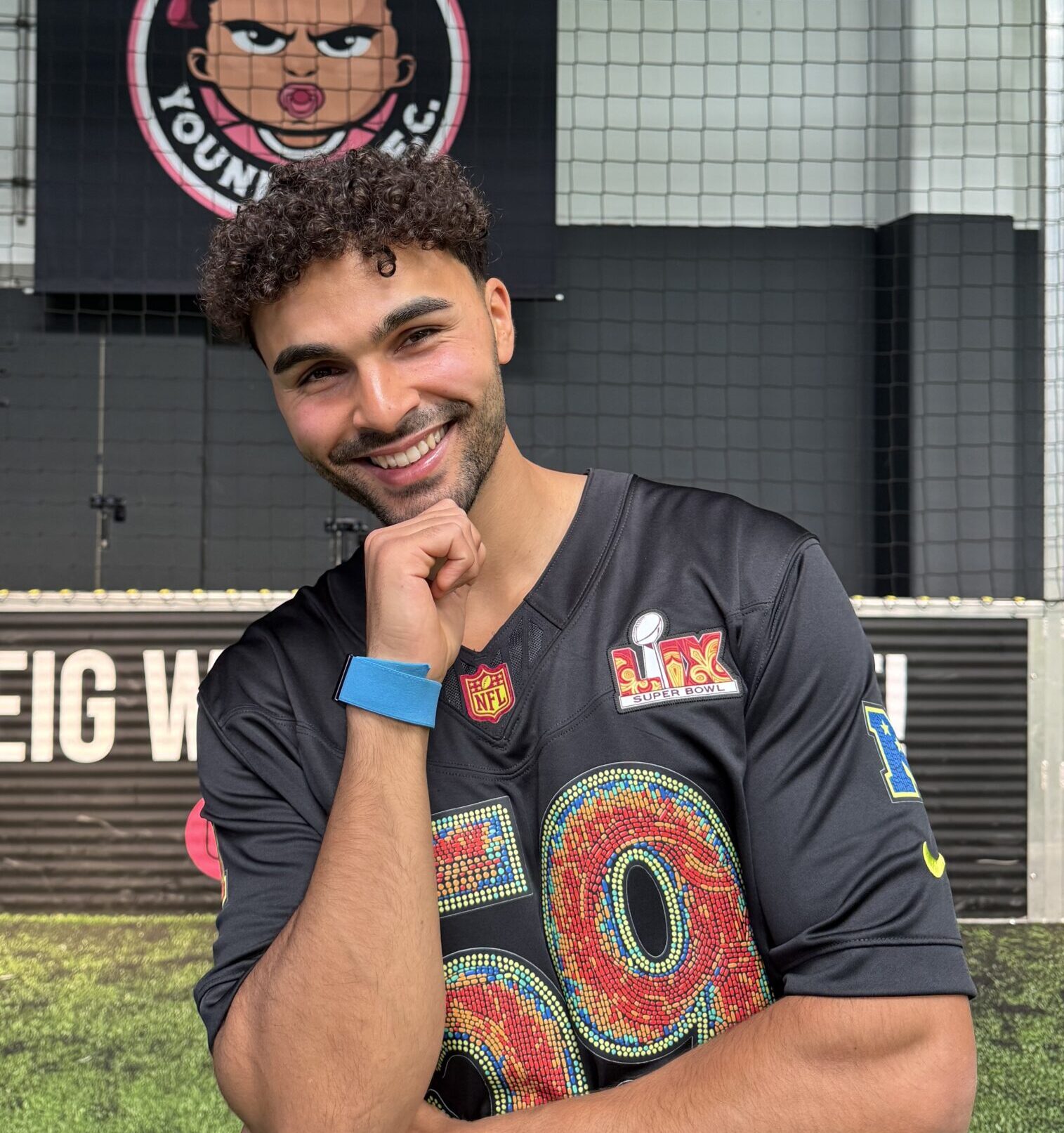 Younes Zarou, lächelnd porträtiert, trägt ein dunkelblaues Trikot mit bunten Applikationen und NFL-Logos.