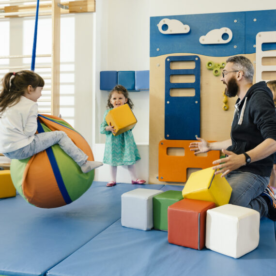Ein männlicher Erzieher sitzt auf einer blauen Matte in einem hellen Turnraum und spielt mit drei Kindern. Ein Mädchen sitzt auf einem bunten Sitzball, ein anderes hält einen gelben Baustein hoch und ein drittes Kind steht daneben. Im Hintergrund sind eine Kletterwand und verschiedene Turngeräte zu sehen.