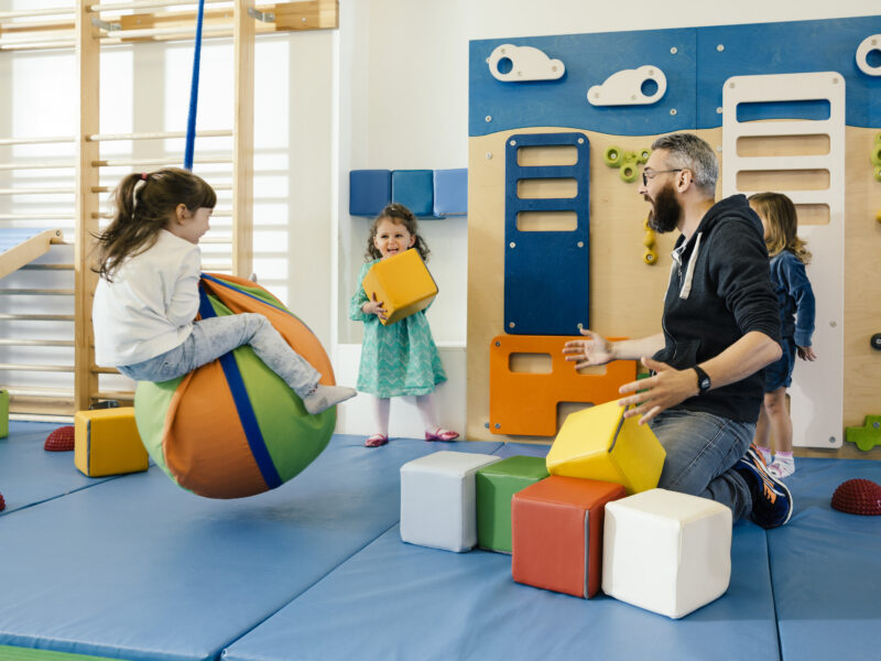 Ein männlicher Erzieher sitzt auf einer blauen Matte in einem hellen Turnraum und spielt mit drei Kindern. Ein Mädchen sitzt auf einem bunten Sitzball, ein anderes hält einen gelben Baustein hoch und ein drittes Kind steht daneben. Im Hintergrund sind eine Kletterwand und verschiedene Turngeräte zu sehen.