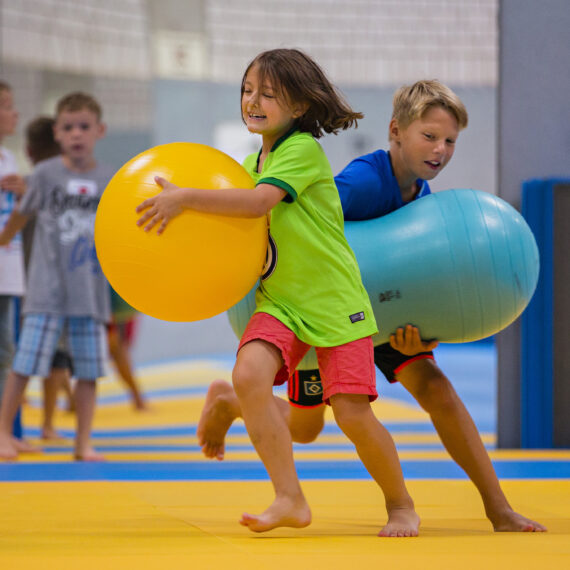 Zwei Kinder, ein Mädchen in grünem T-Shirt und ein Junge in blauem T-Shirt, tragen große Bälle barfuß auf einer gelben Matte. Im Hintergrund sind weitere Kinder zu sehen, die ebenfalls aktiv sind.