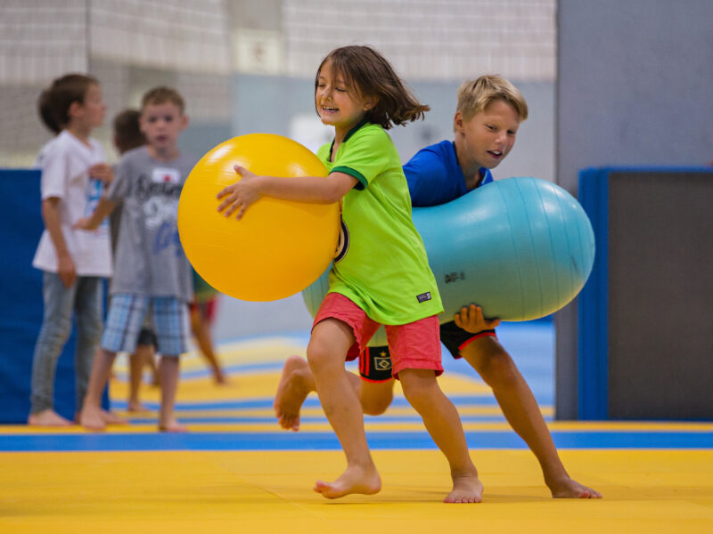 Zwei Kinder, ein Mädchen in grünem T-Shirt und ein Junge in blauem T-Shirt, tragen große Bälle barfuß auf einer gelben Matte. Im Hintergrund sind weitere Kinder zu sehen, die ebenfalls aktiv sind.