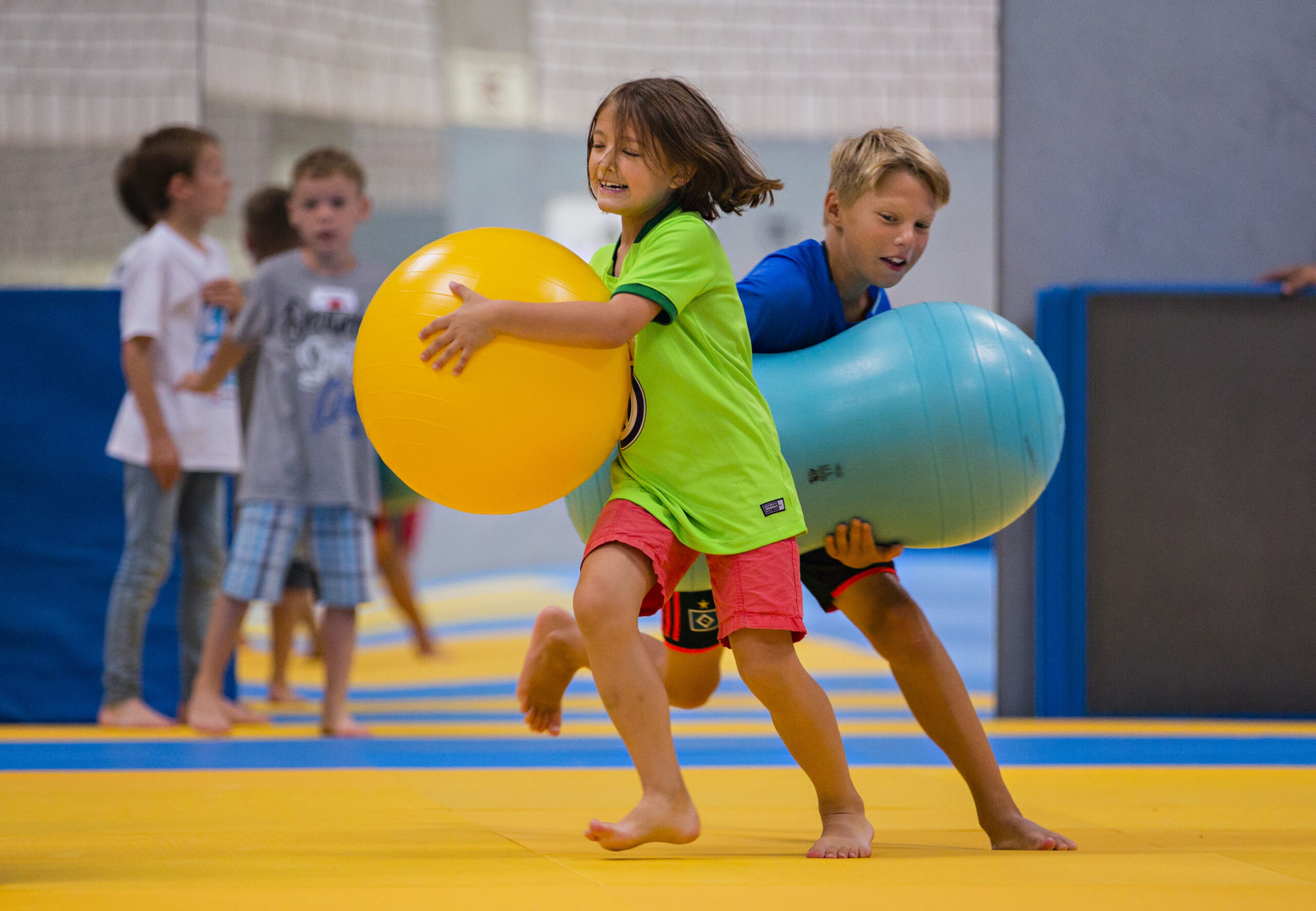 Zwei Kinder, ein Mädchen in grünem T-Shirt und ein Junge in blauem T-Shirt, tragen große Bälle barfuß auf einer gelben Matte. Im Hintergrund sind weitere Kinder zu sehen, die ebenfalls aktiv sind.