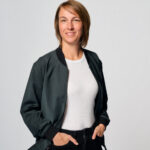 Eine Frau mit kurzen, hellbraunen Haaren lächelt in die Kamera. Sie trägt eine schwarze Jacke über einem weißen T-Shirt und dunkle Hosen. Ihre Hände stecken in den Hosentaschen.