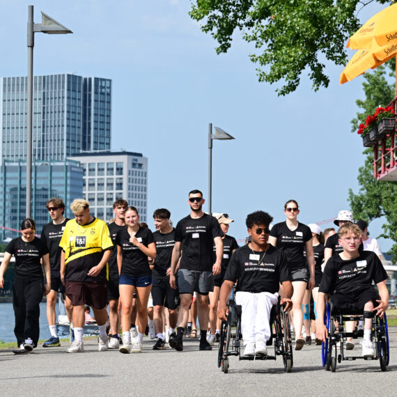 Eine Gruppe von jungen Menschen, darunter auch Rollstuhlfahrer, schiebt oder begleitet Rollstühle auf einem Gehweg entlang eines Flusses. Alle tragen dunkle T-Shirts mit dem Aufdruck der DFL Stiftung. Im Hintergrund ist die Skyline von Frankfurt zu sehen.