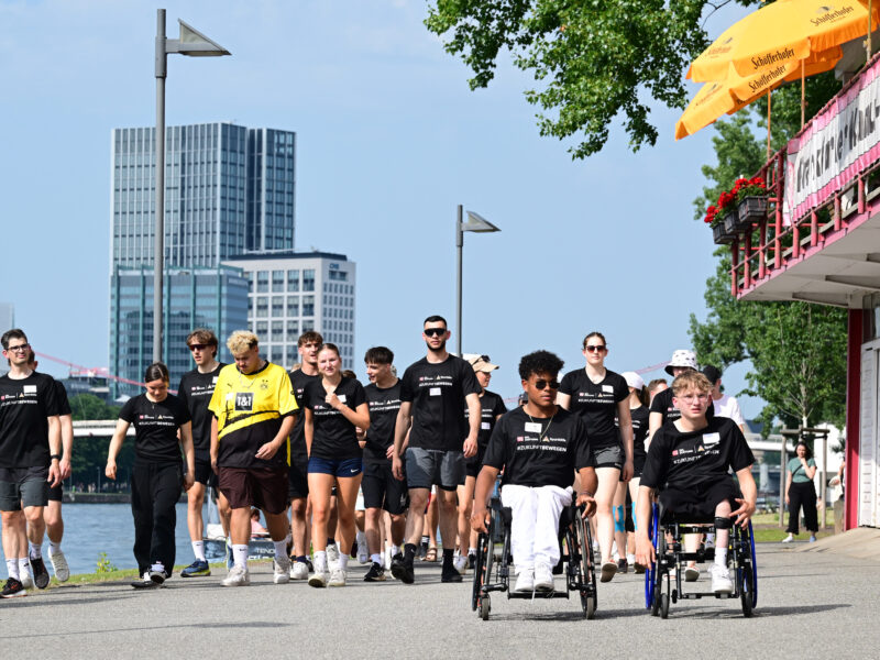 Eine Gruppe von jungen Menschen, darunter auch Rollstuhlfahrer, schiebt oder begleitet Rollstühle auf einem Gehweg entlang eines Flusses. Alle tragen dunkle T-Shirts mit dem Aufdruck der DFL Stiftung. Im Hintergrund ist die Skyline von Frankfurt zu sehen.
