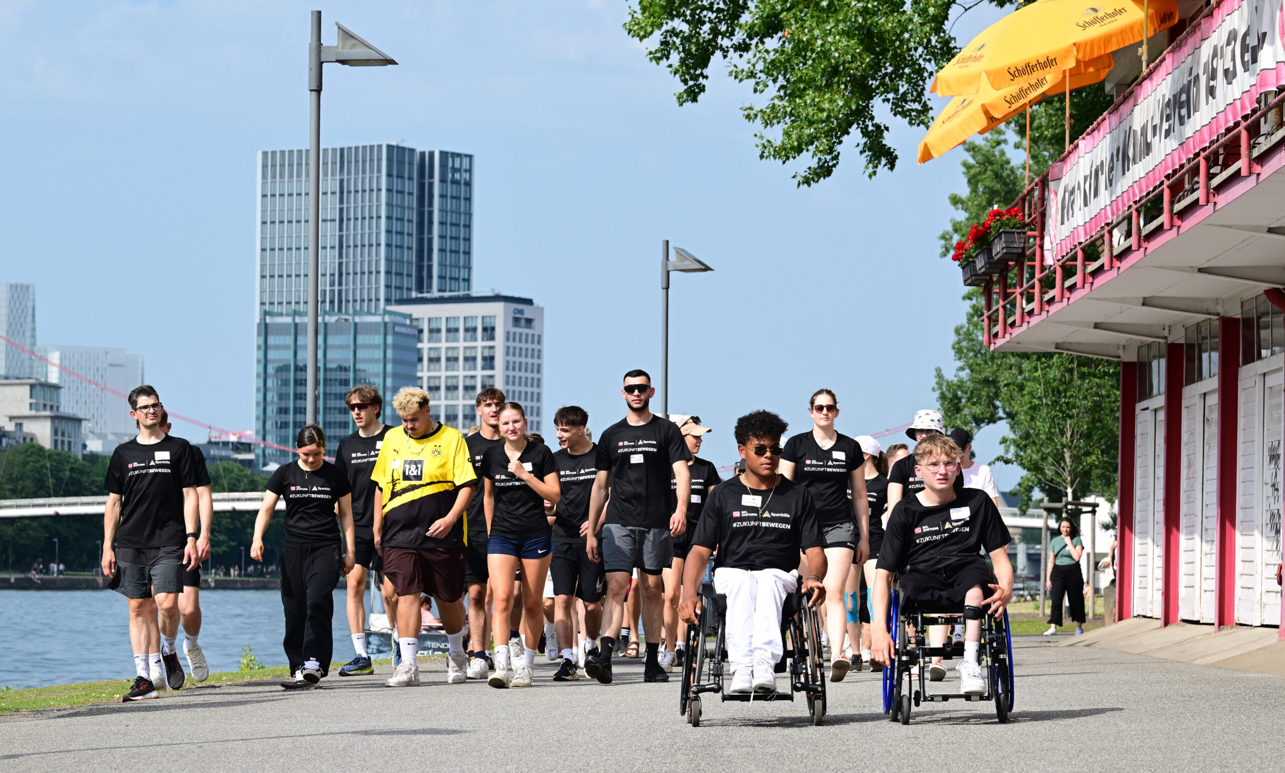 Eine Gruppe von jungen Menschen, darunter auch Rollstuhlfahrer, schiebt oder begleitet Rollstühle auf einem Gehweg entlang eines Flusses. Alle tragen dunkle T-Shirts mit dem Aufdruck der DFL Stiftung. Im Hintergrund ist die Skyline von Frankfurt zu sehen.